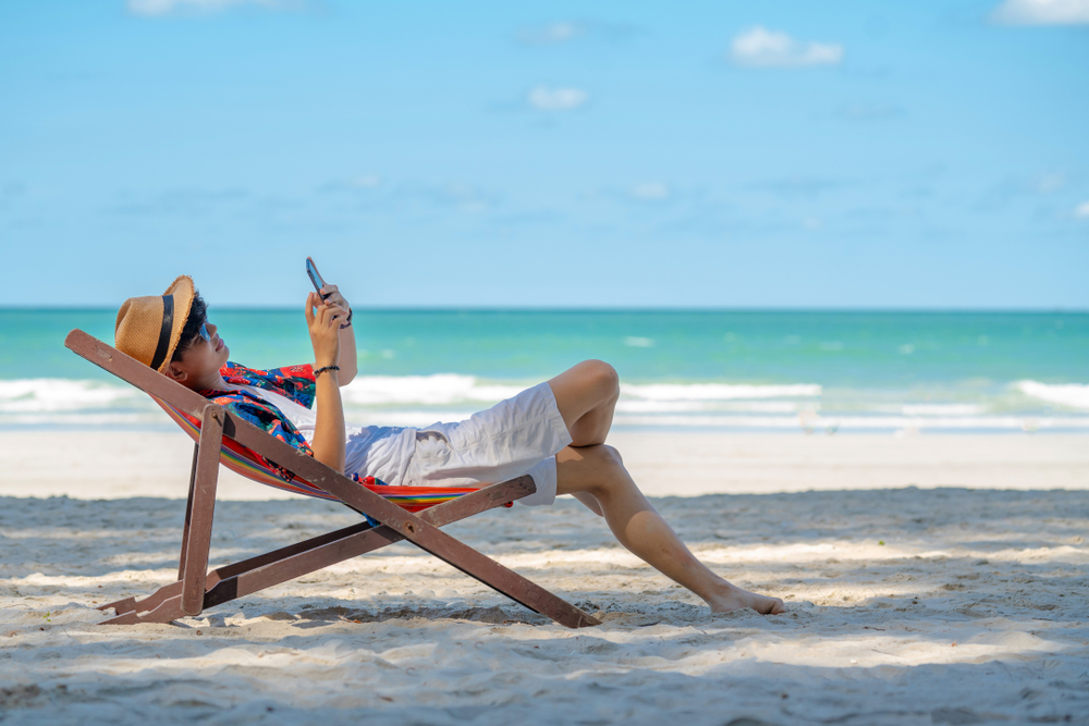 Eine Person hält ein Smartphone in der Hand und liegt am Strand in einer Liege.
